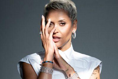 Fado singer Mariza, a Portuguese woman with short bleached blonde hair, wears a white sleeveless shirt against a dark background and gazes towards the camera from behind the fingers of her left hand.